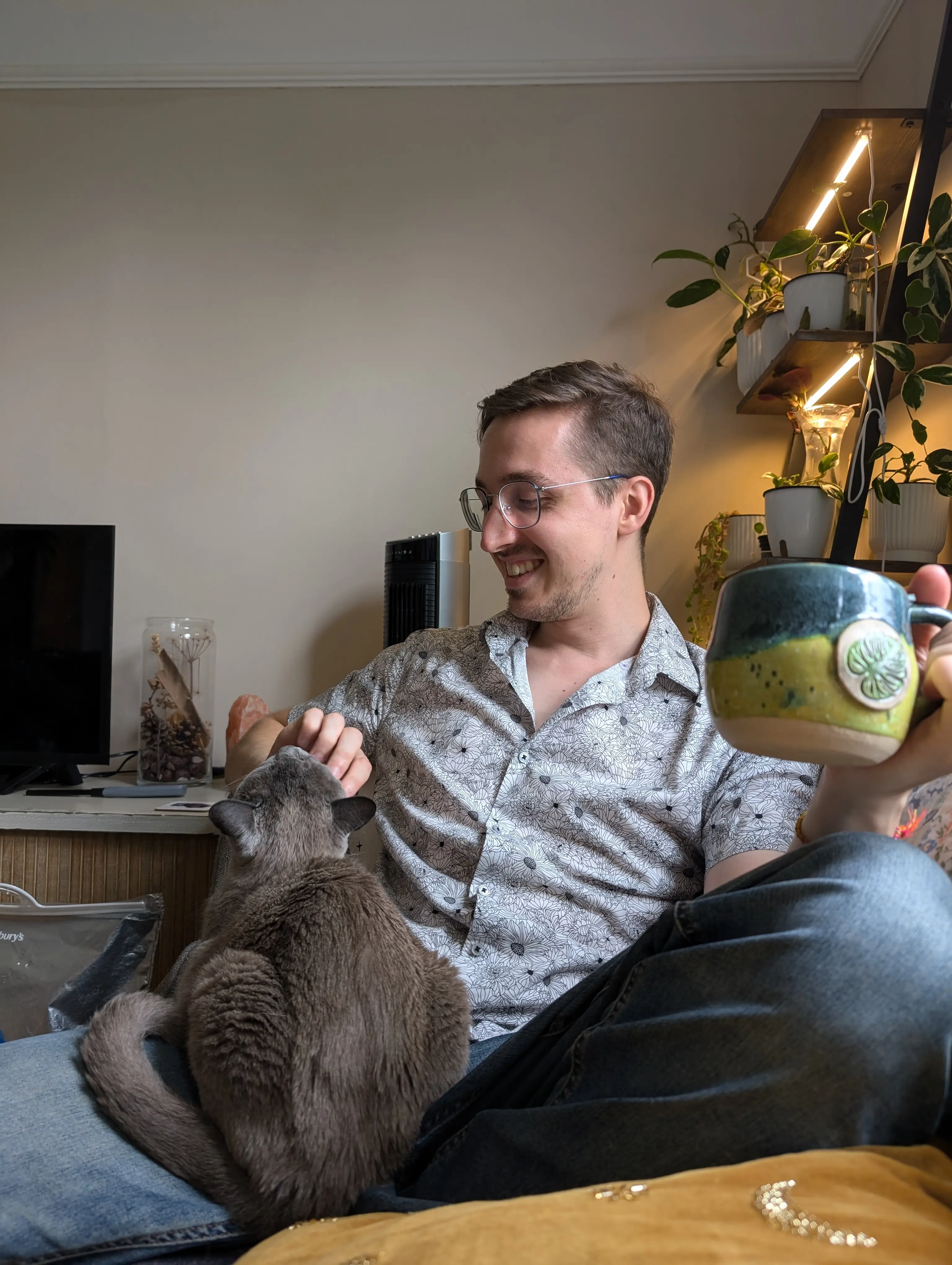 Simon sits on a sofa, coffee mug in one hand, petting a cat on his lap. He wears a black and white floral shirt, blue jeans, blue glasses, and seems to be enjoying the cat's company.